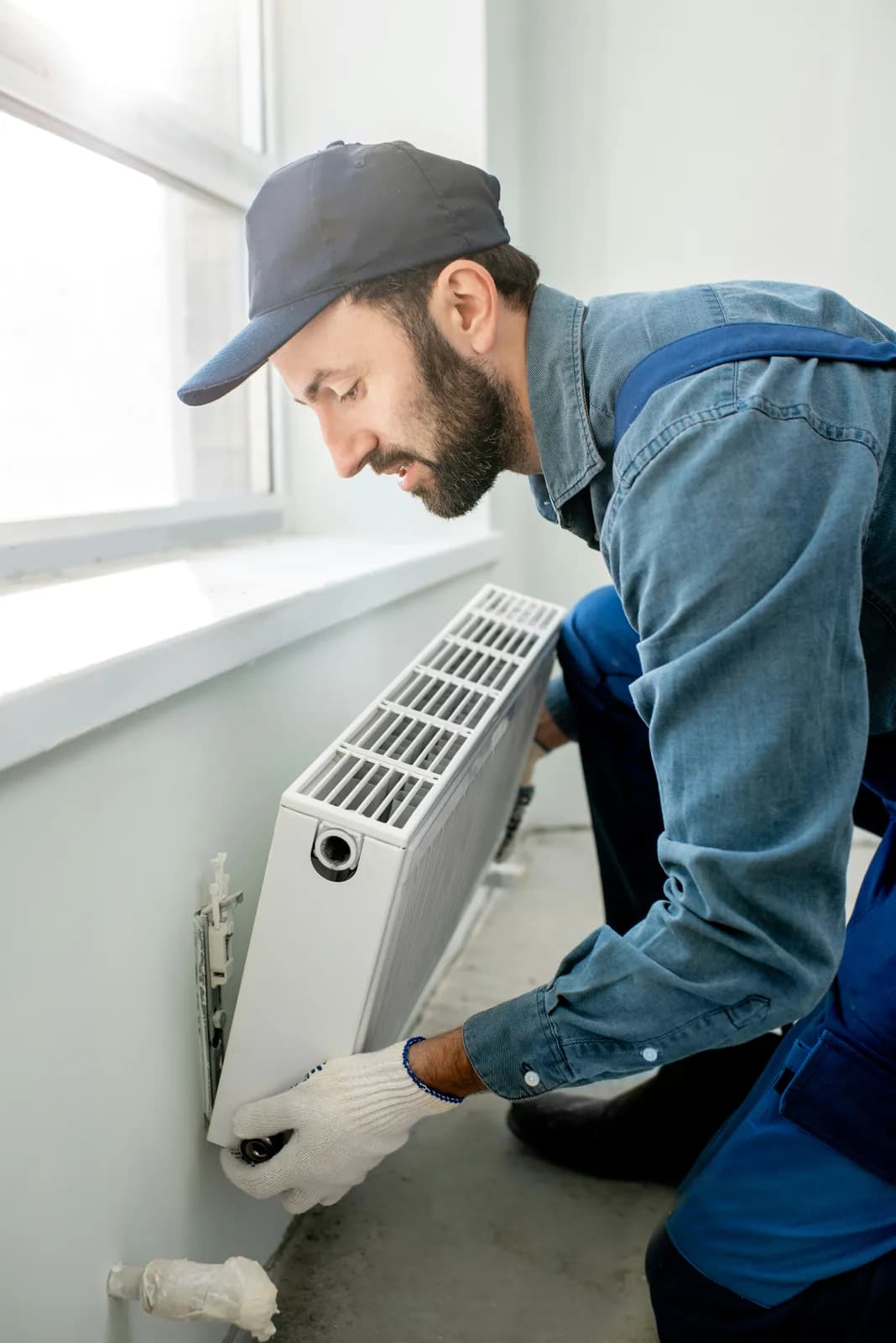 Plombier barbu en casquette et gants blancs ajustant un radiateur blanc sous une fenêtre