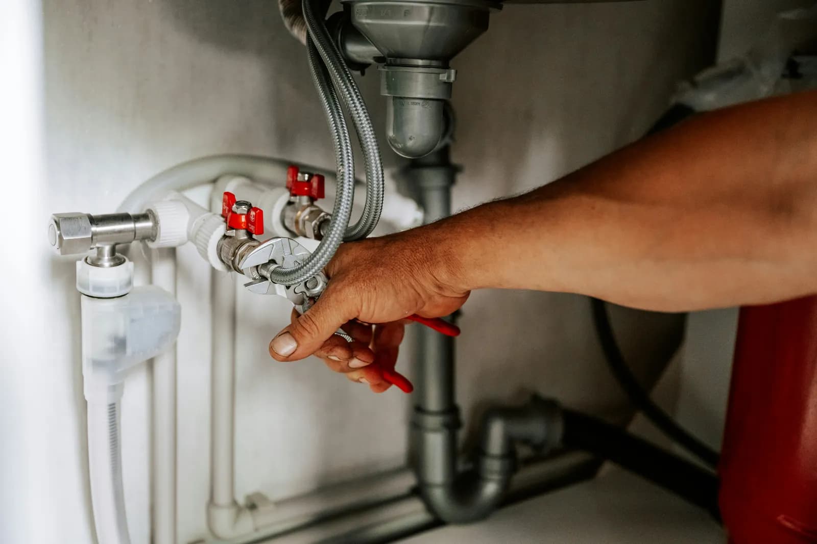 Main serrant à la pince multiprise un raccord de flexible sous un évier, près des vannes rouges et du siphon